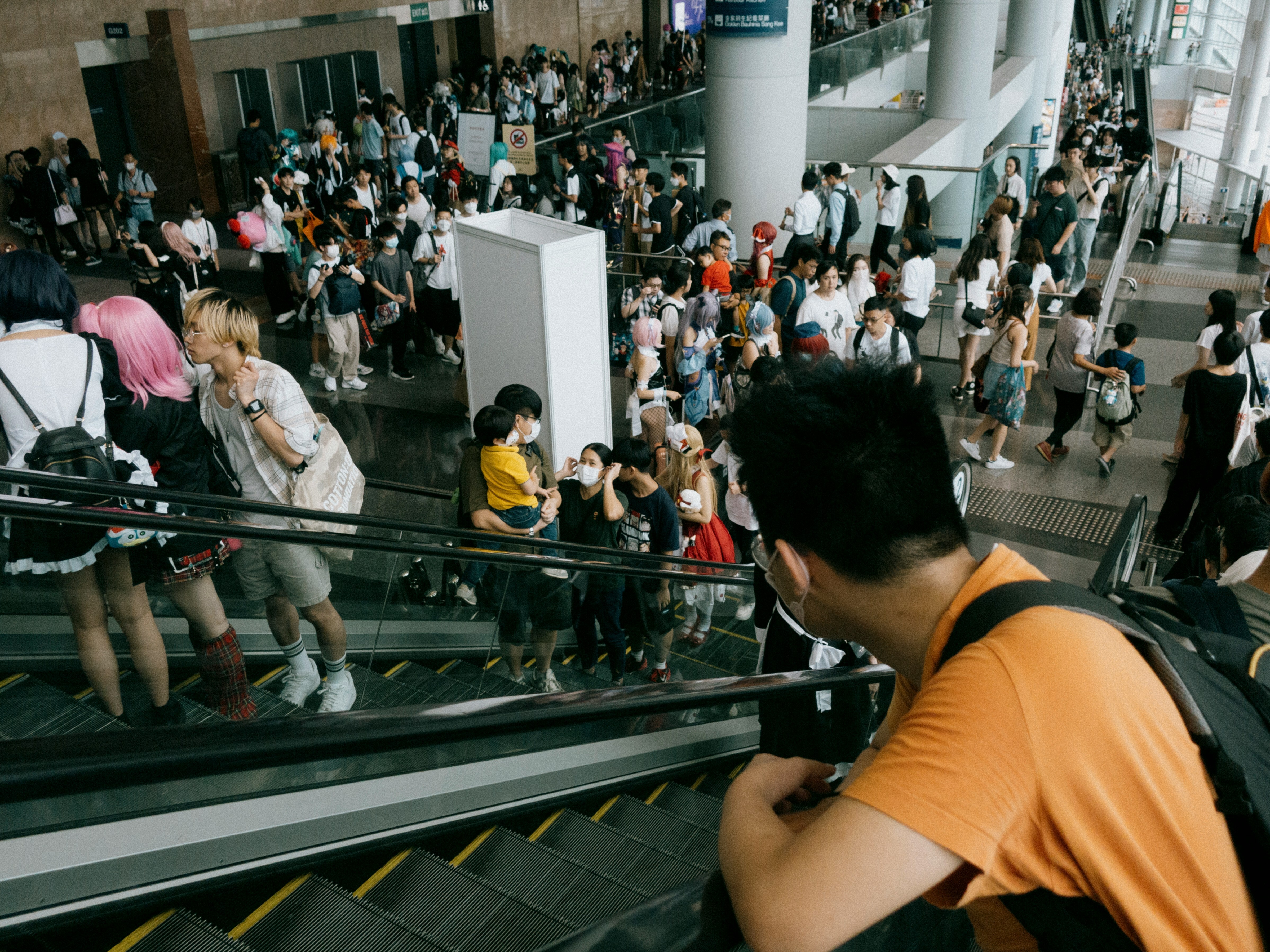 a convention at the HKCEC, a man in an orange shirt is going up the escalator looking at a crowd of people