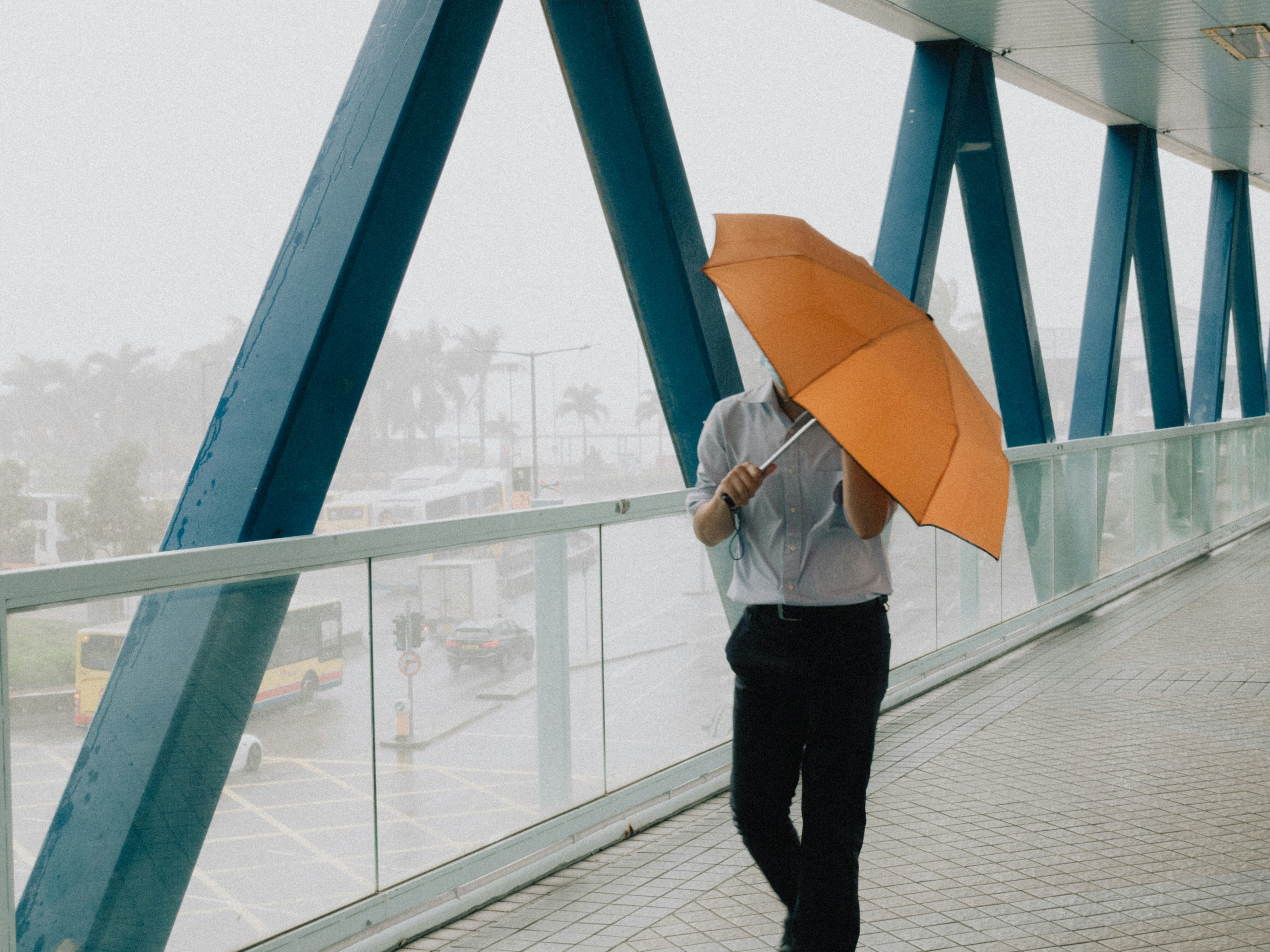 A man hold an orange umbrella while walking along an overpass where it is raining