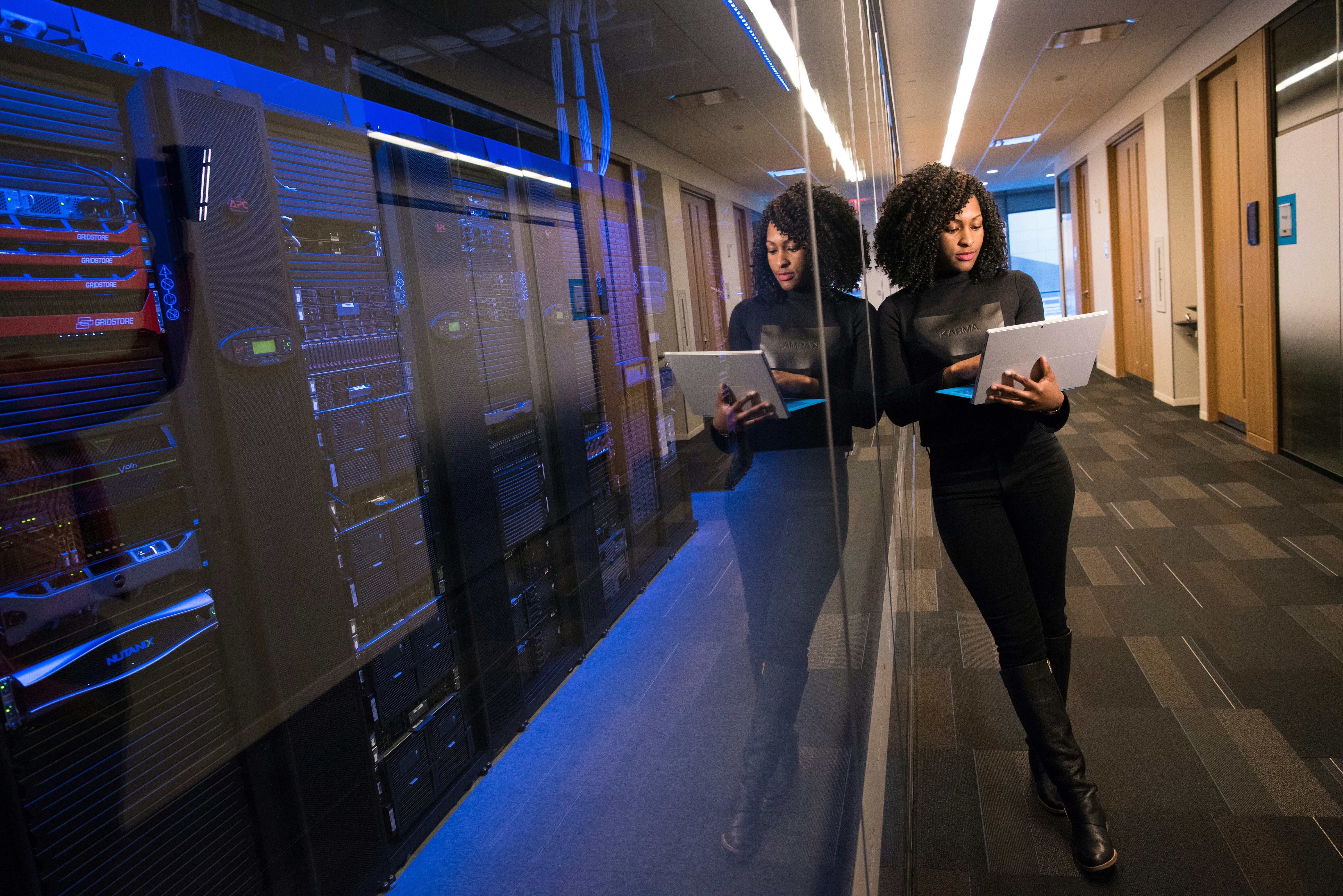 A worker on a laptop leaning on the wall next to a rack of servers